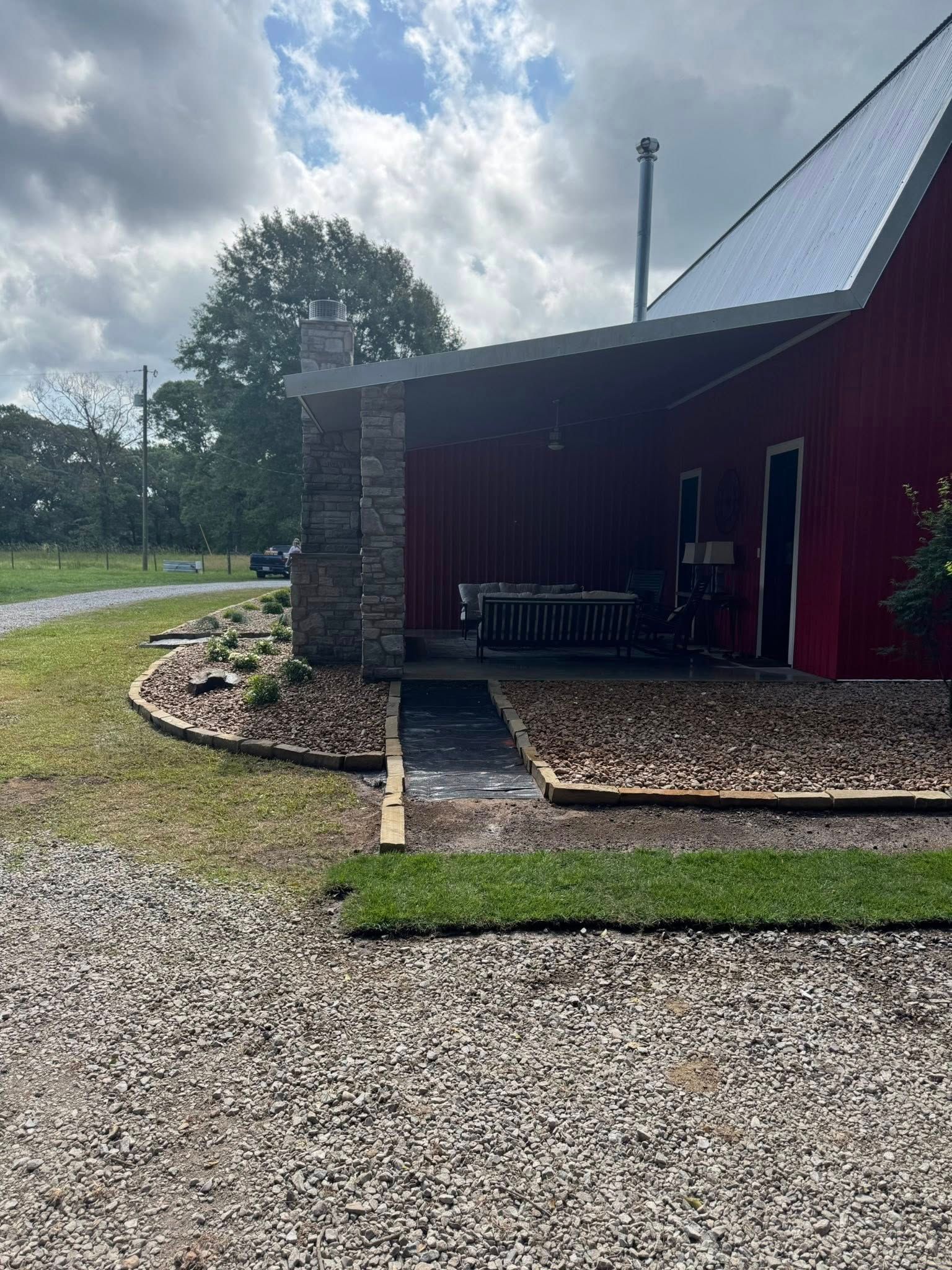 Red building with covered porch and stone chimney, gravel path, and grass under cloudy sky.