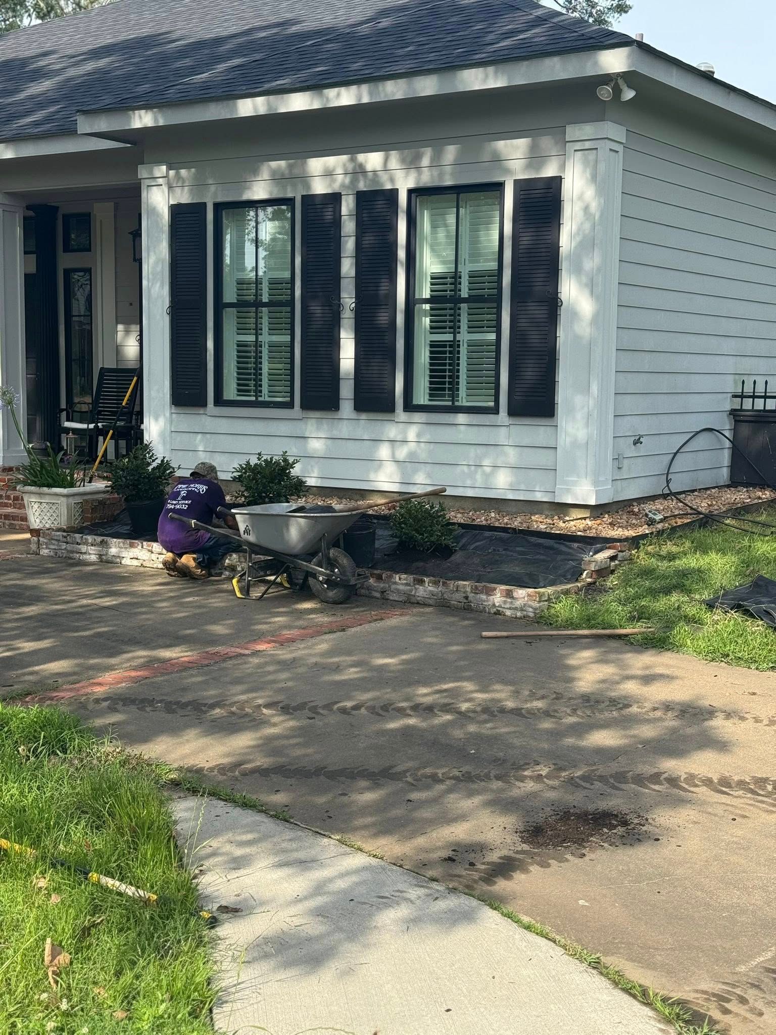 Person gardening in front of a white house with black shutters; driveway in foreground.