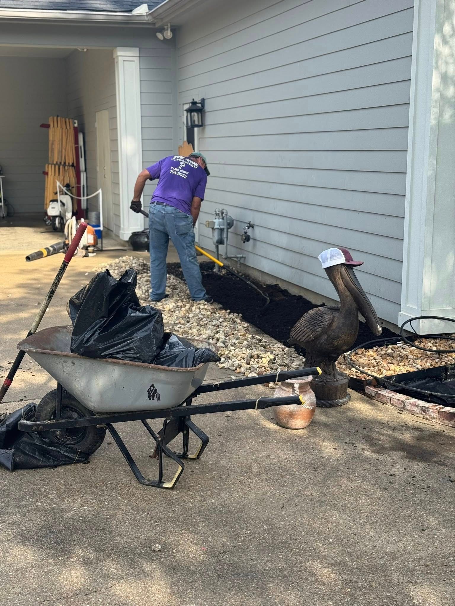 Man digging in a garden bed next to a house; wheelbarrow and tools nearby.