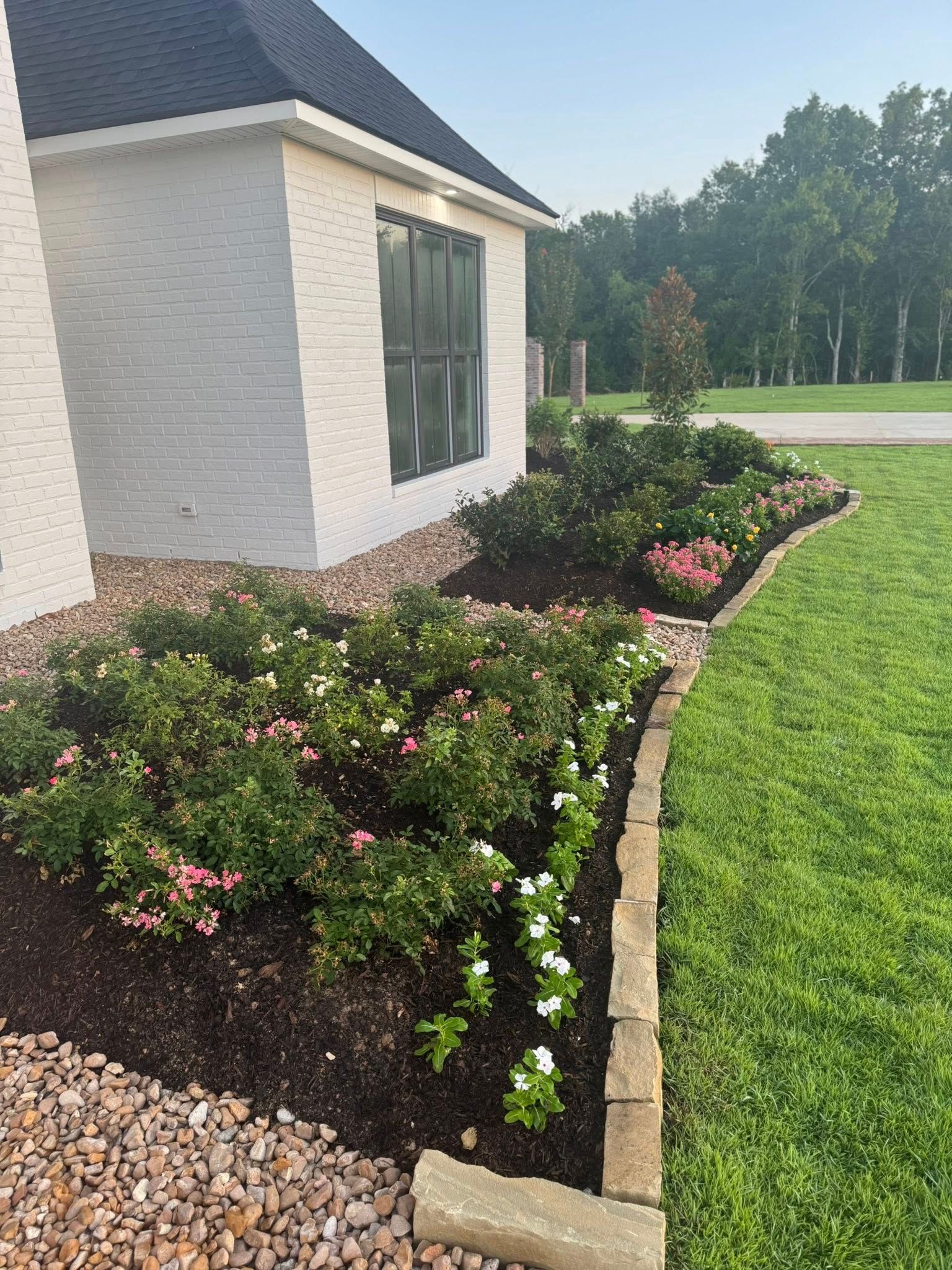 Flowerbed in front of a white brick house with dark shutters, bordered by stone and green lawn.