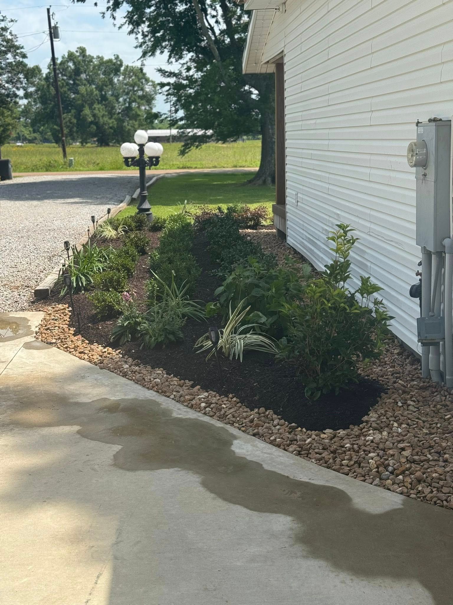 Landscaped bed with plants and gravel border next to a white building. A lamppost stands nearby.