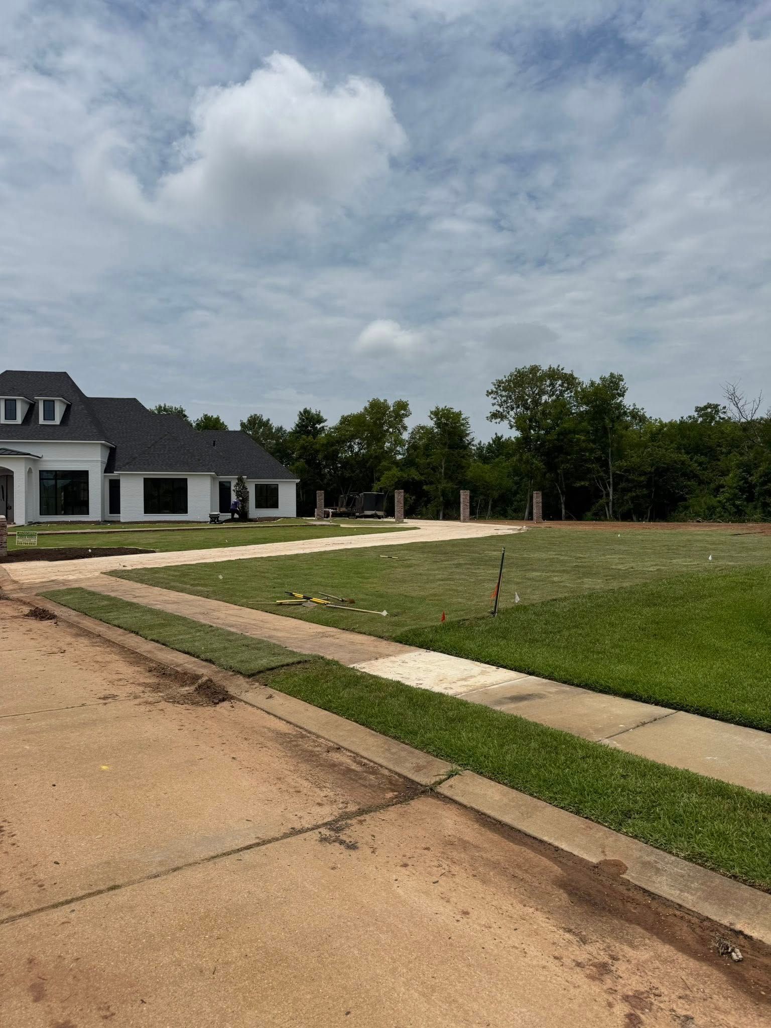 White house with black roof, driveway, and newly planted grass under cloudy sky.