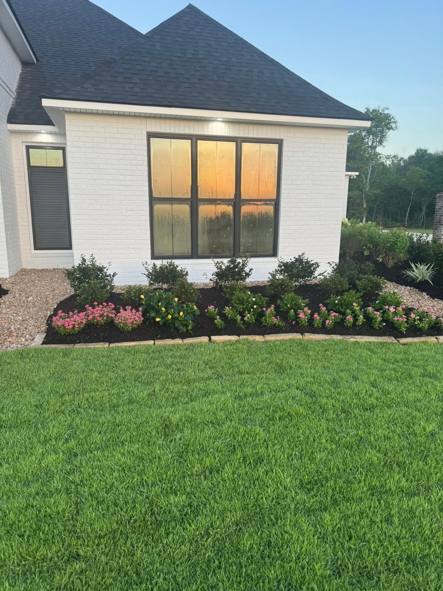 White brick house with a flower bed, green lawn, and sunset reflected in windows.