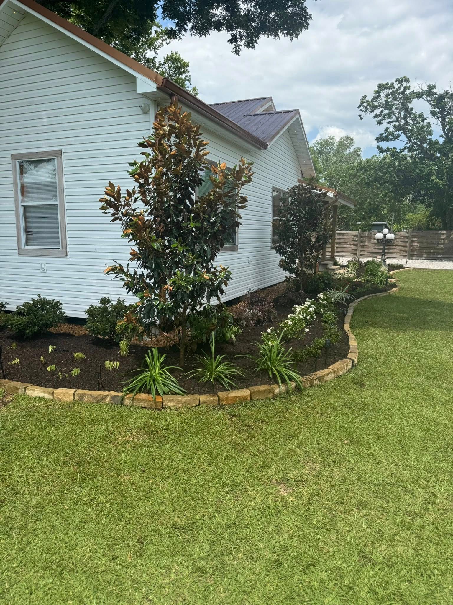 White house with a brick-edged flower bed. Trees and greenery are next to the house, with green grass in the foreground.