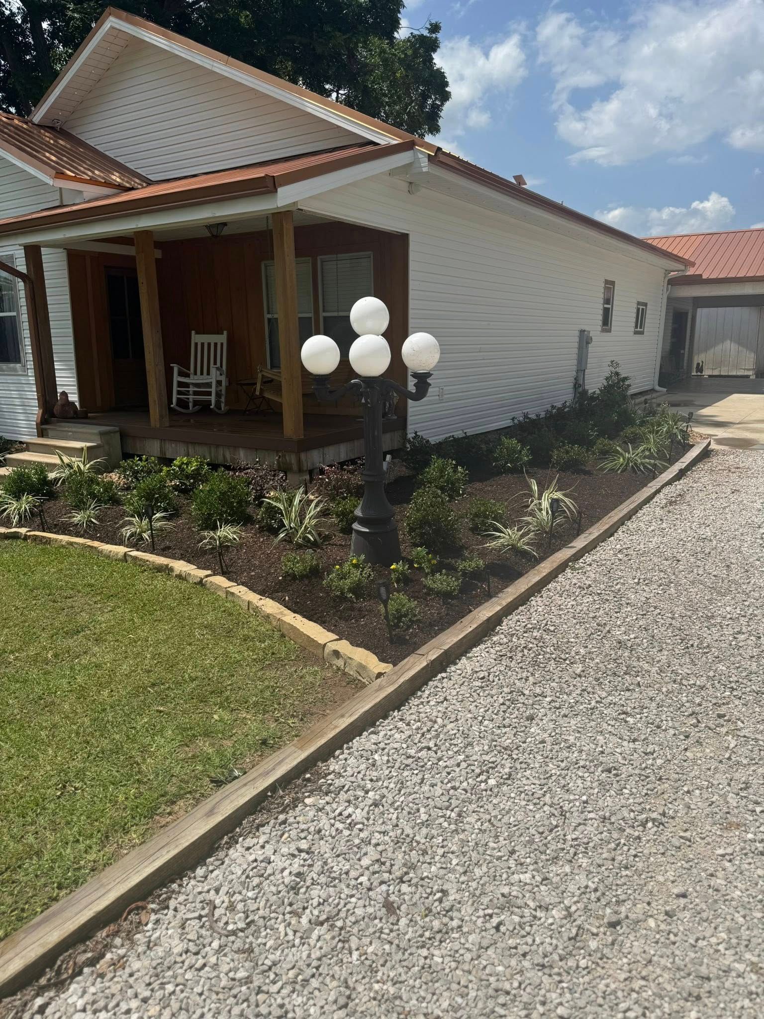White house with porch and gravel driveway, landscaped flower bed in front.