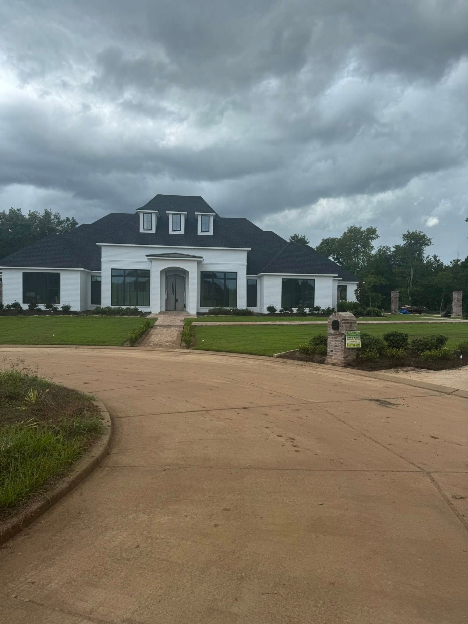 White house with black roof, under a cloudy sky. Long driveway with green grass.