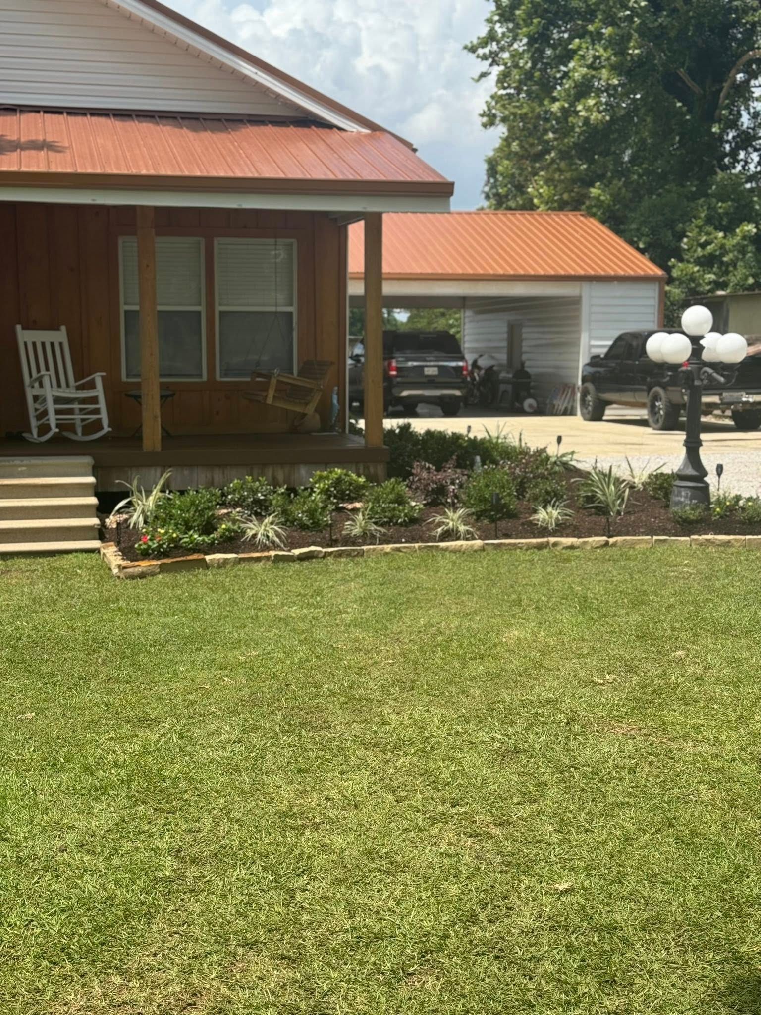 A house with a porch and a garden; an open carport with vehicles is in the background.