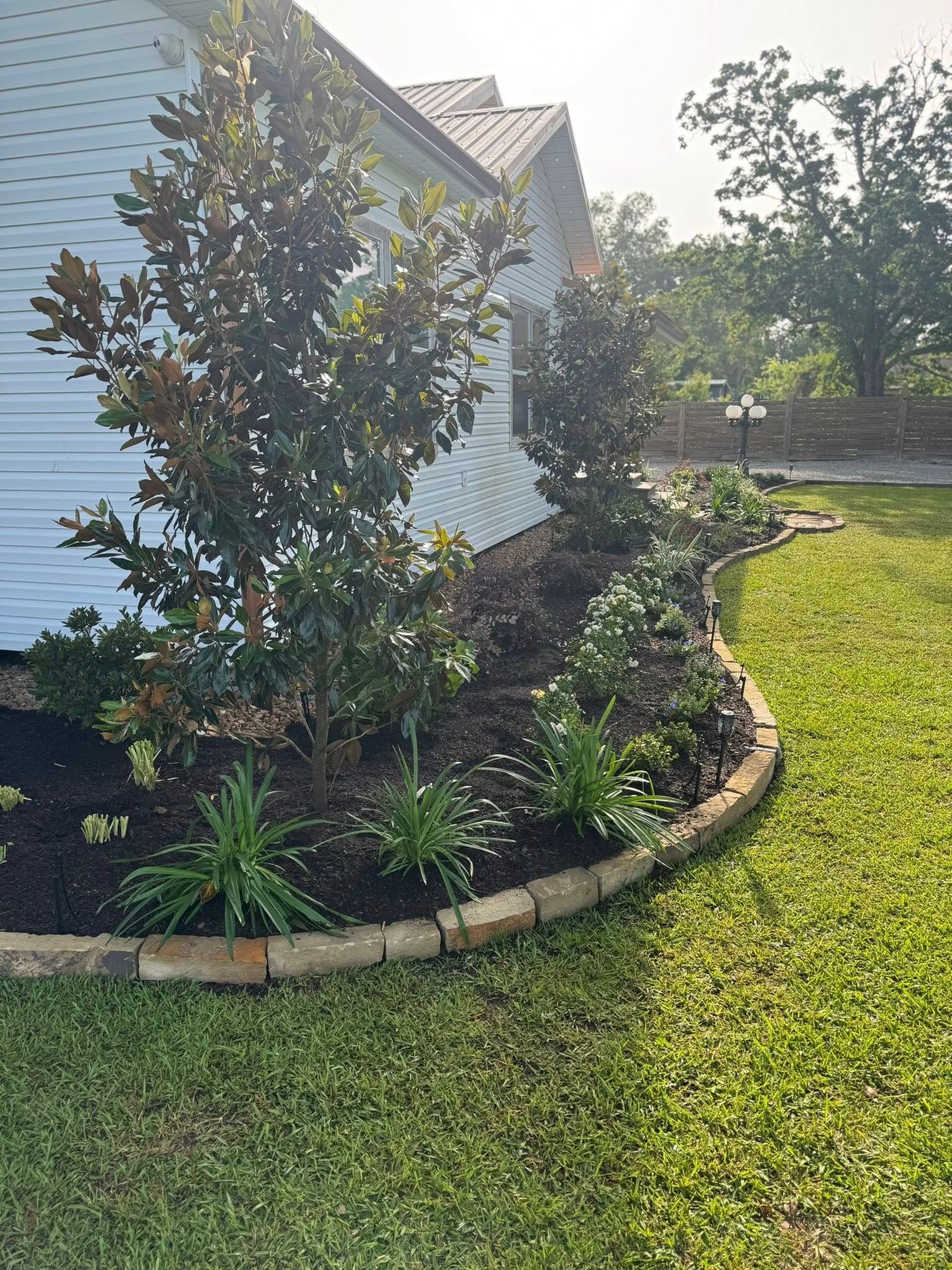 A garden bed with a curved brick border, planted with various green plants and small trees, next to a white house.