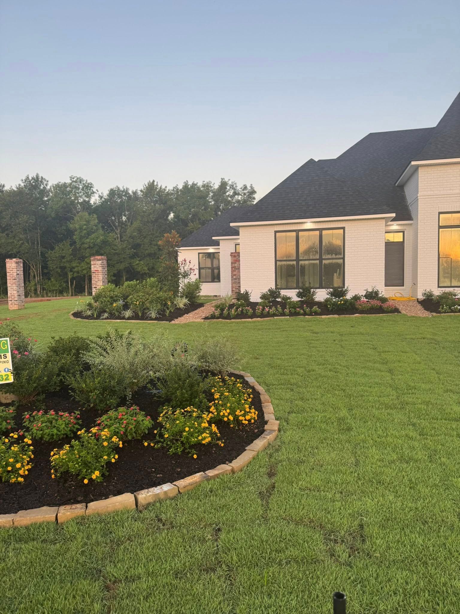 A house with a manicured lawn and flower beds, trees in the background, and a blue sky.