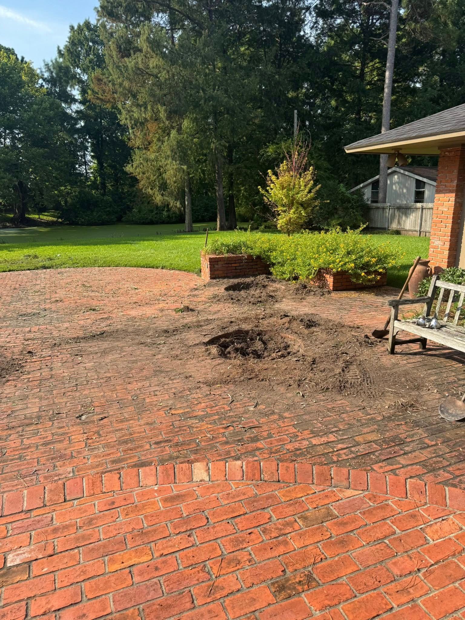 Brick patio with dark spots, two planters, and a bench; trees and grass in the background.