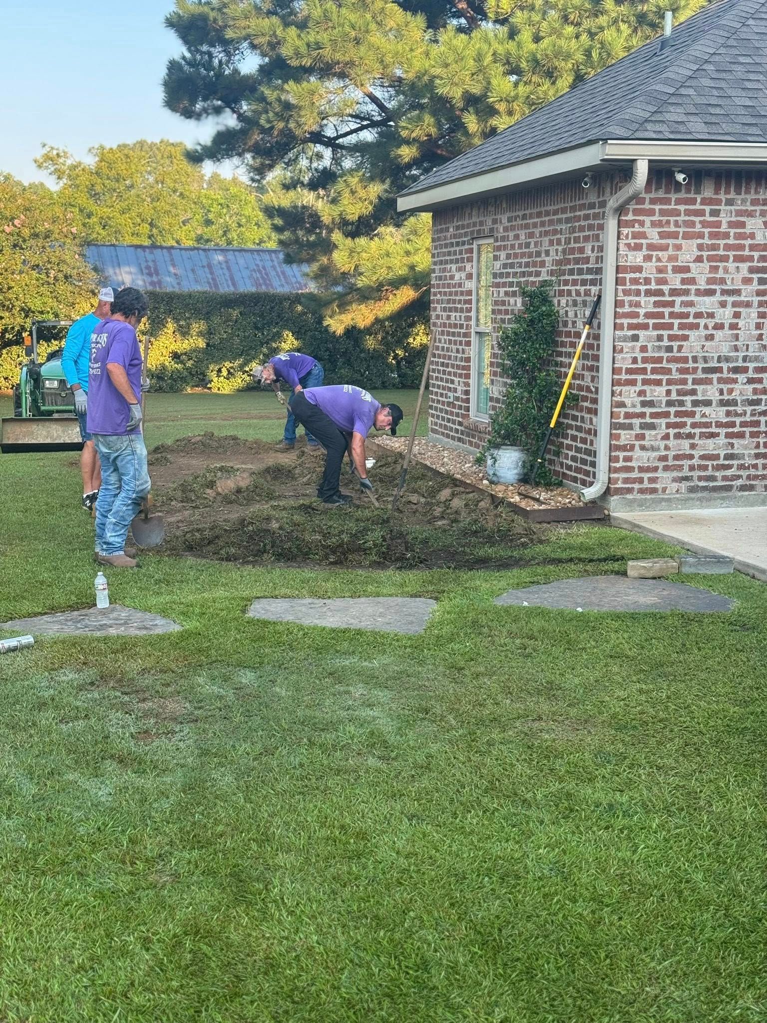 People in purple shirts digging in dirt near a brick house. Green grass in the foreground.