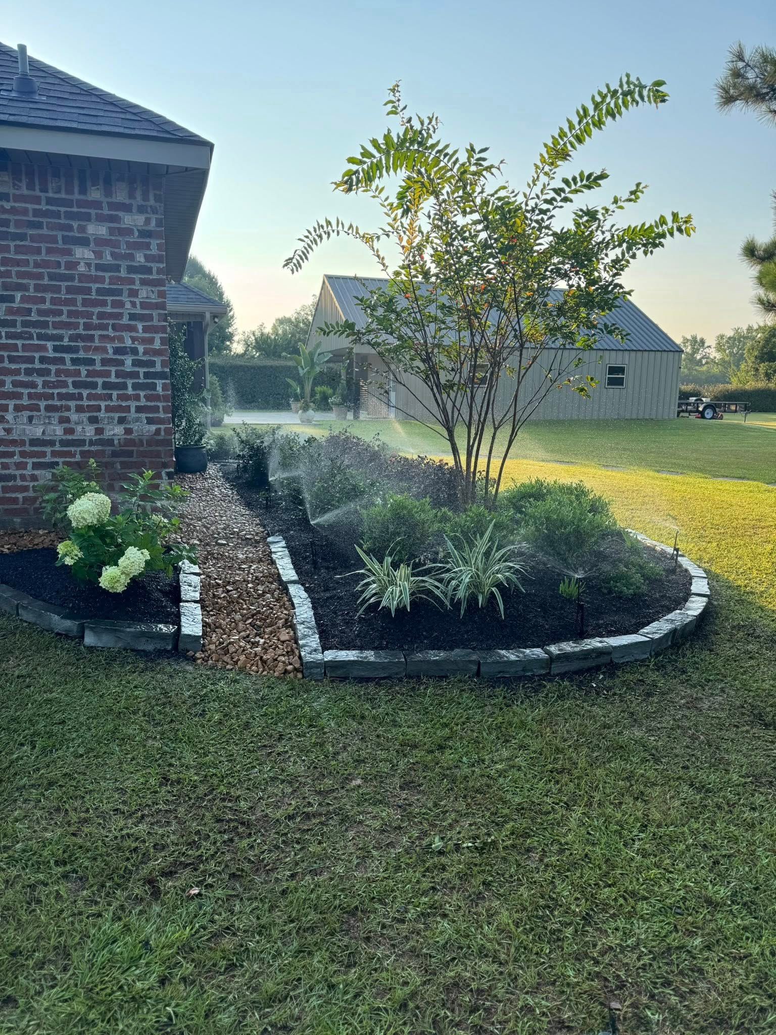 Garden bed with a variety of plants, bordered by bricks, next to a brick house on a sunny day.