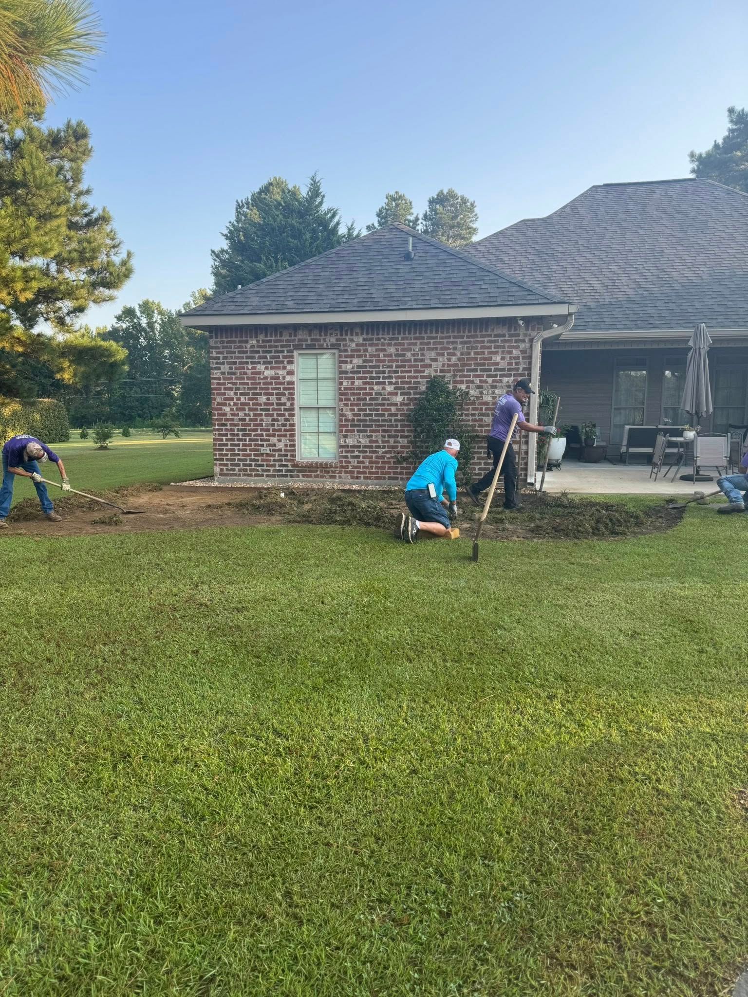 People digging and working in yard next to a brick building on a sunny day.