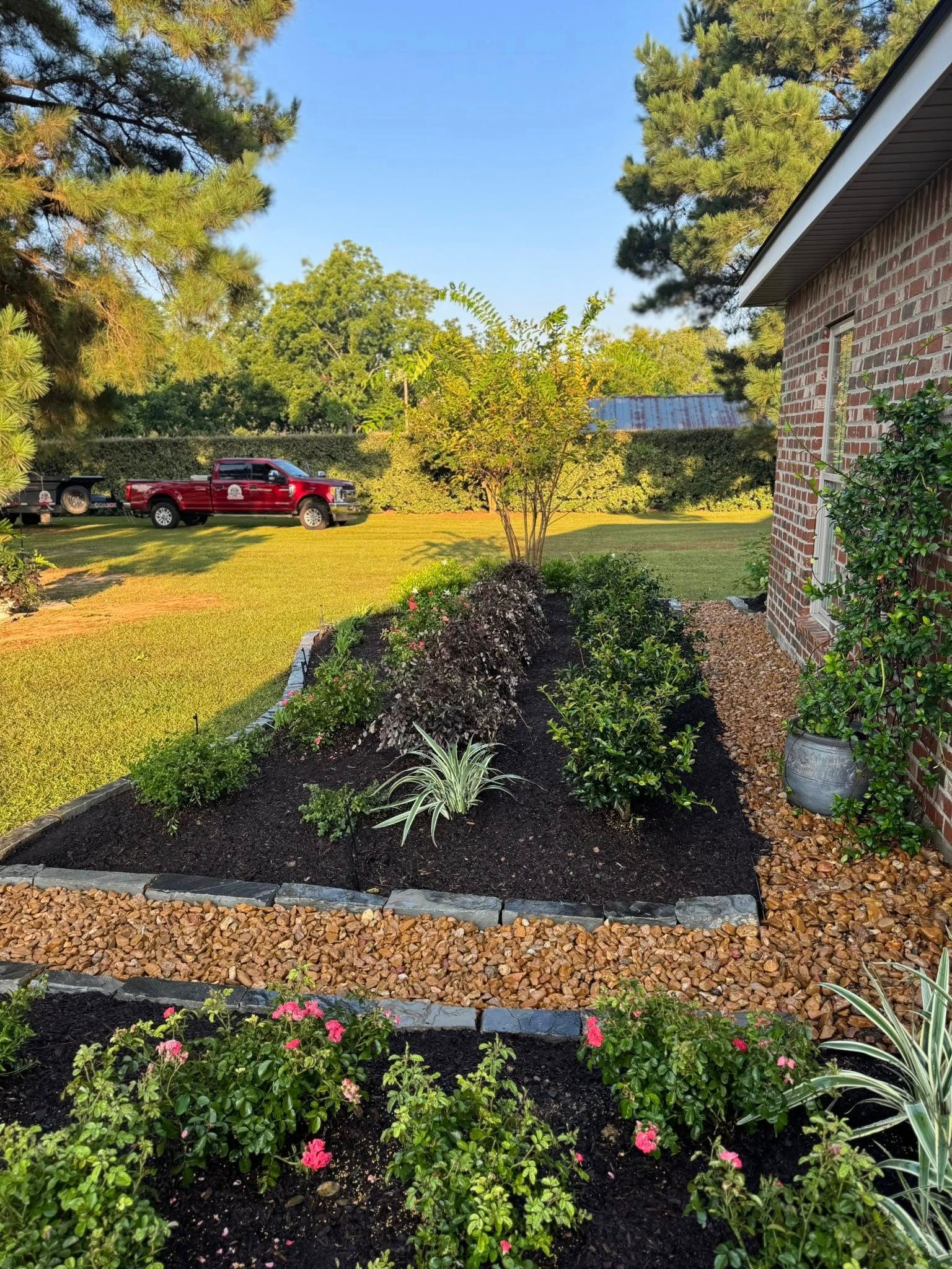 Flower bed next to a brick house, featuring various plants, mulch, and landscaping rocks. A red truck is parked in the background.