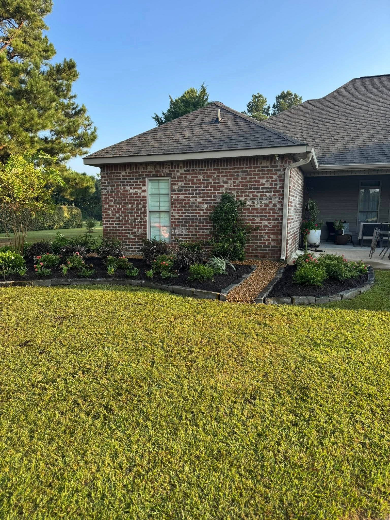 Brick outbuilding with window, surrounded by flowerbeds and green lawn. Adjacent to house with dark roof.