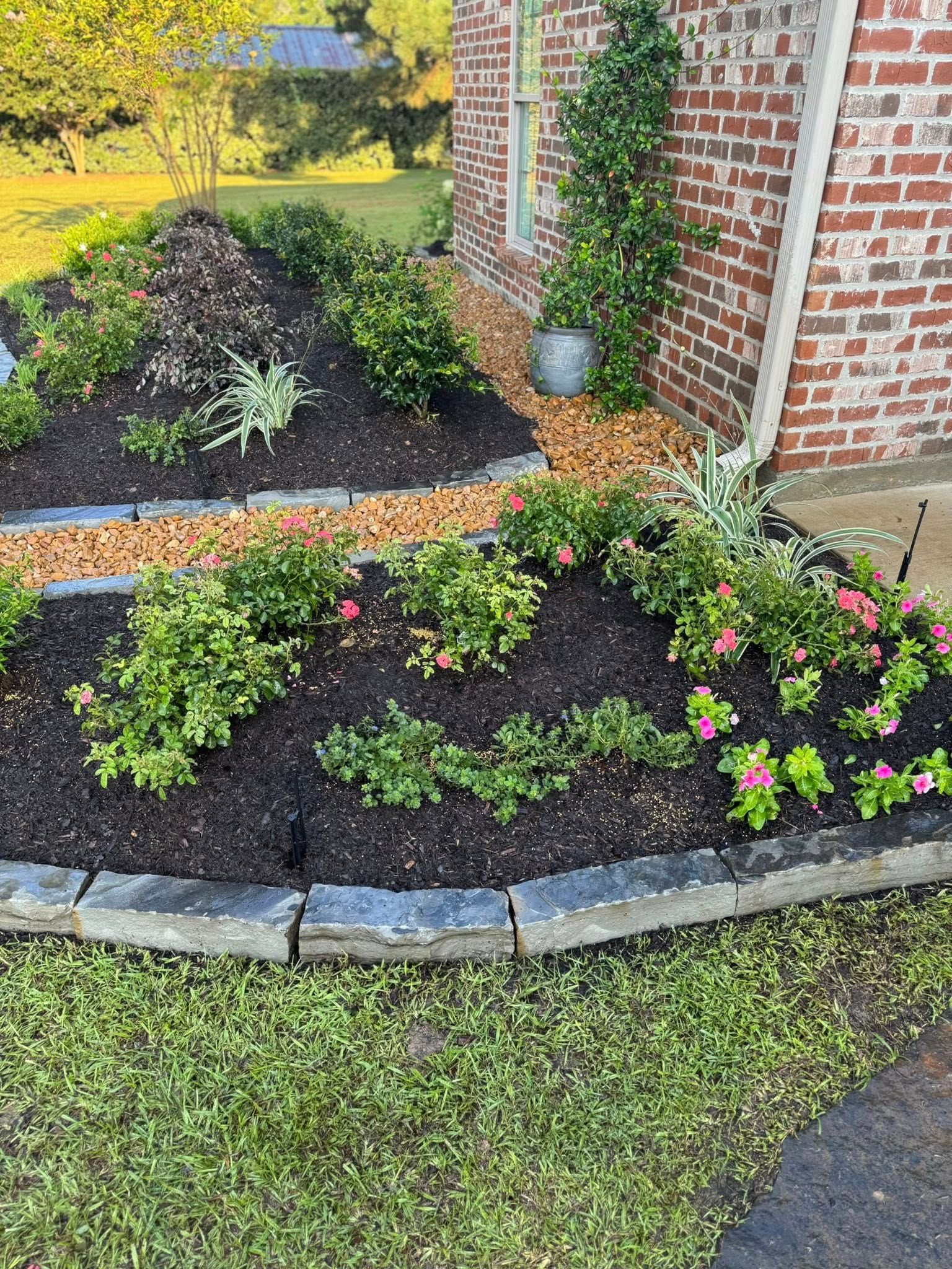 Flowerbed with green plants and pink flowers, bordered by stone and grass, next to a brick house.