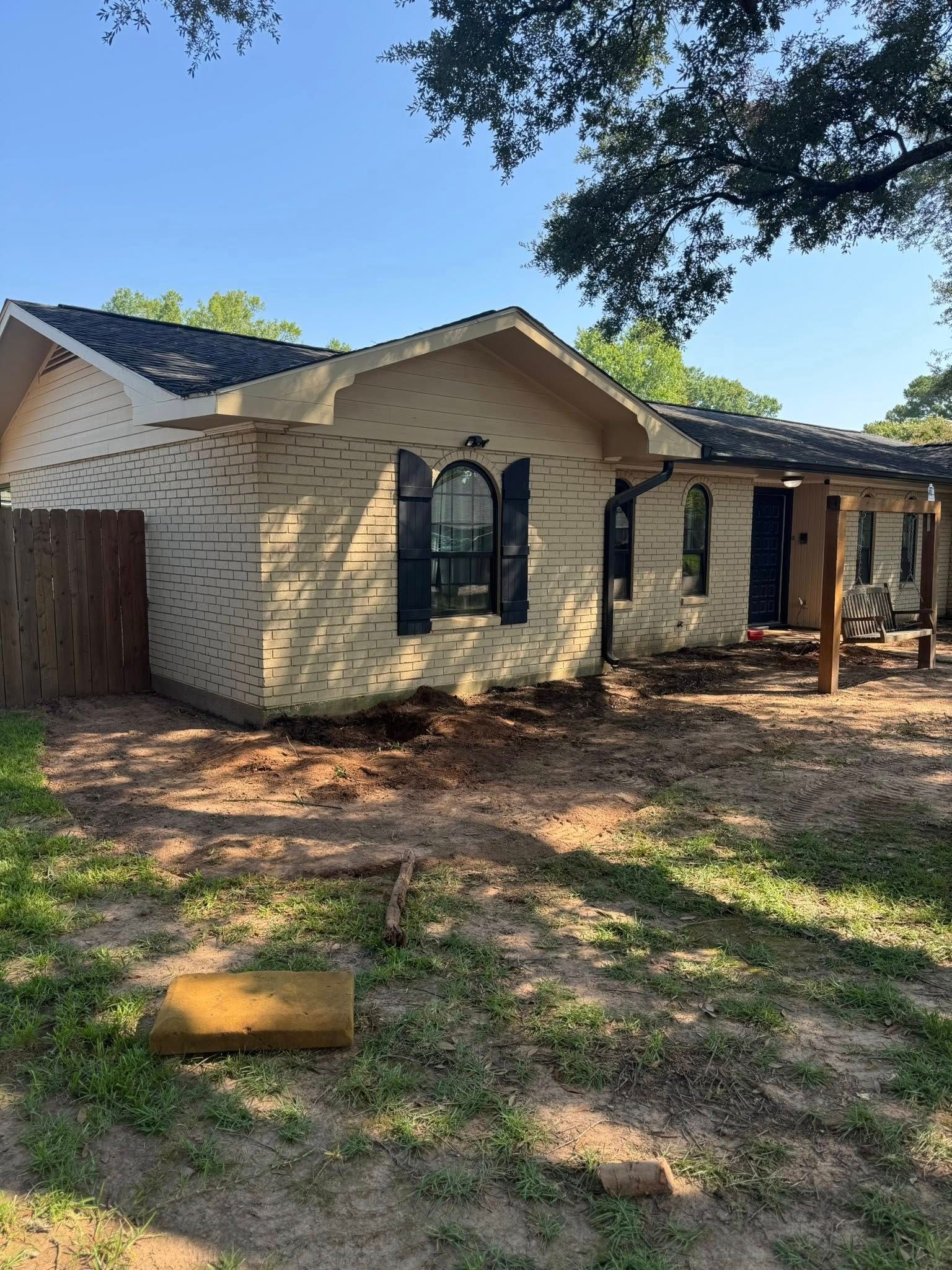 Tan brick house with black shutters, wooden fence, and porch with wooden posts, under a blue sky.