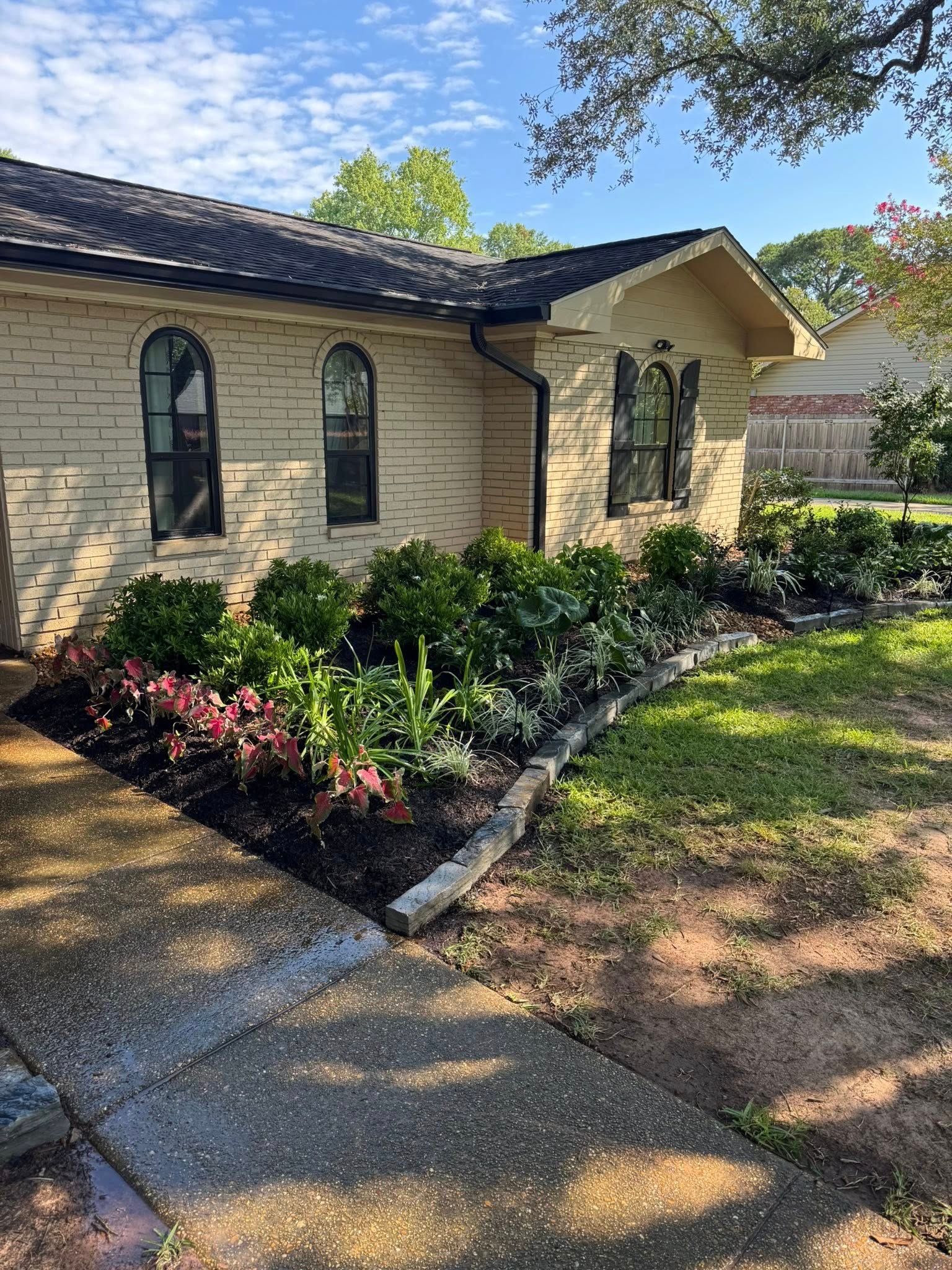 A brick house with a well-maintained flowerbed in front. Dark mulch, green and red plants, and a gray sidewalk.