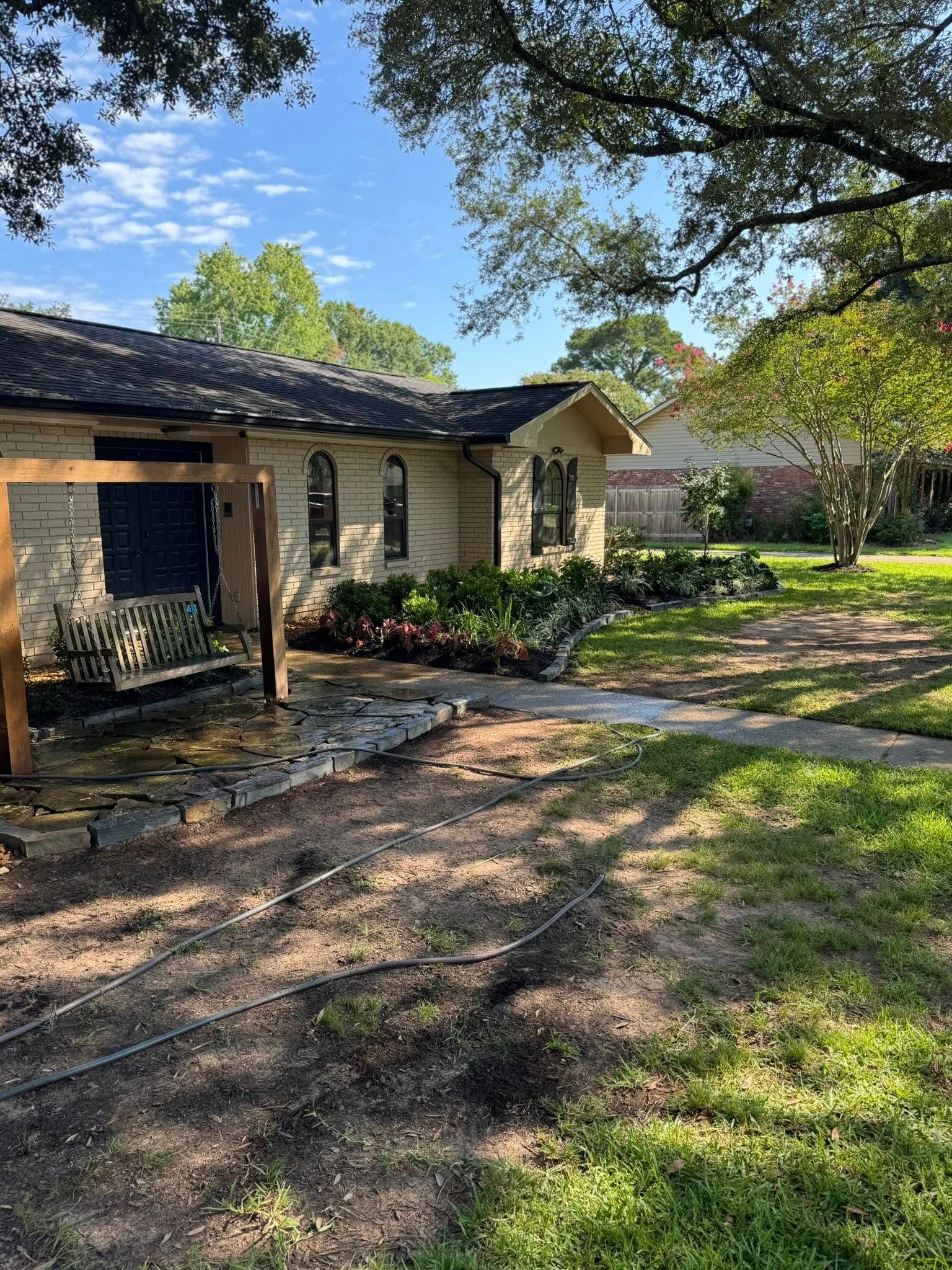 Single-story beige house with dark roof and green yard. Sunny day, trees, pathway in front.