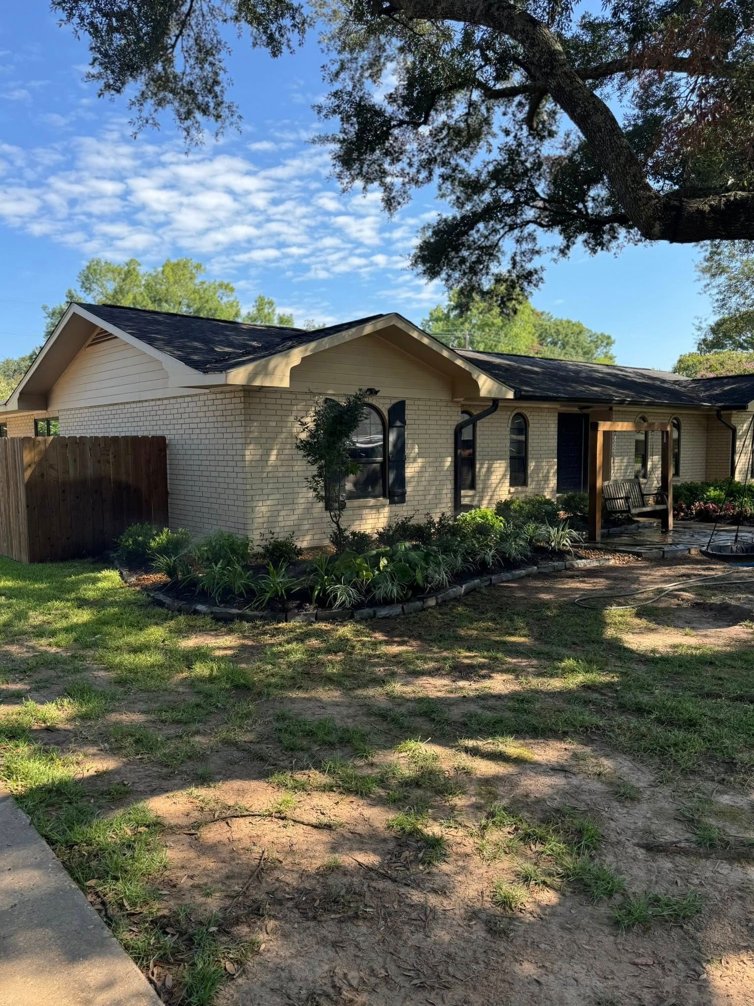 Beige house with a black roof, surrounded by green grass and trees, under a blue sky.