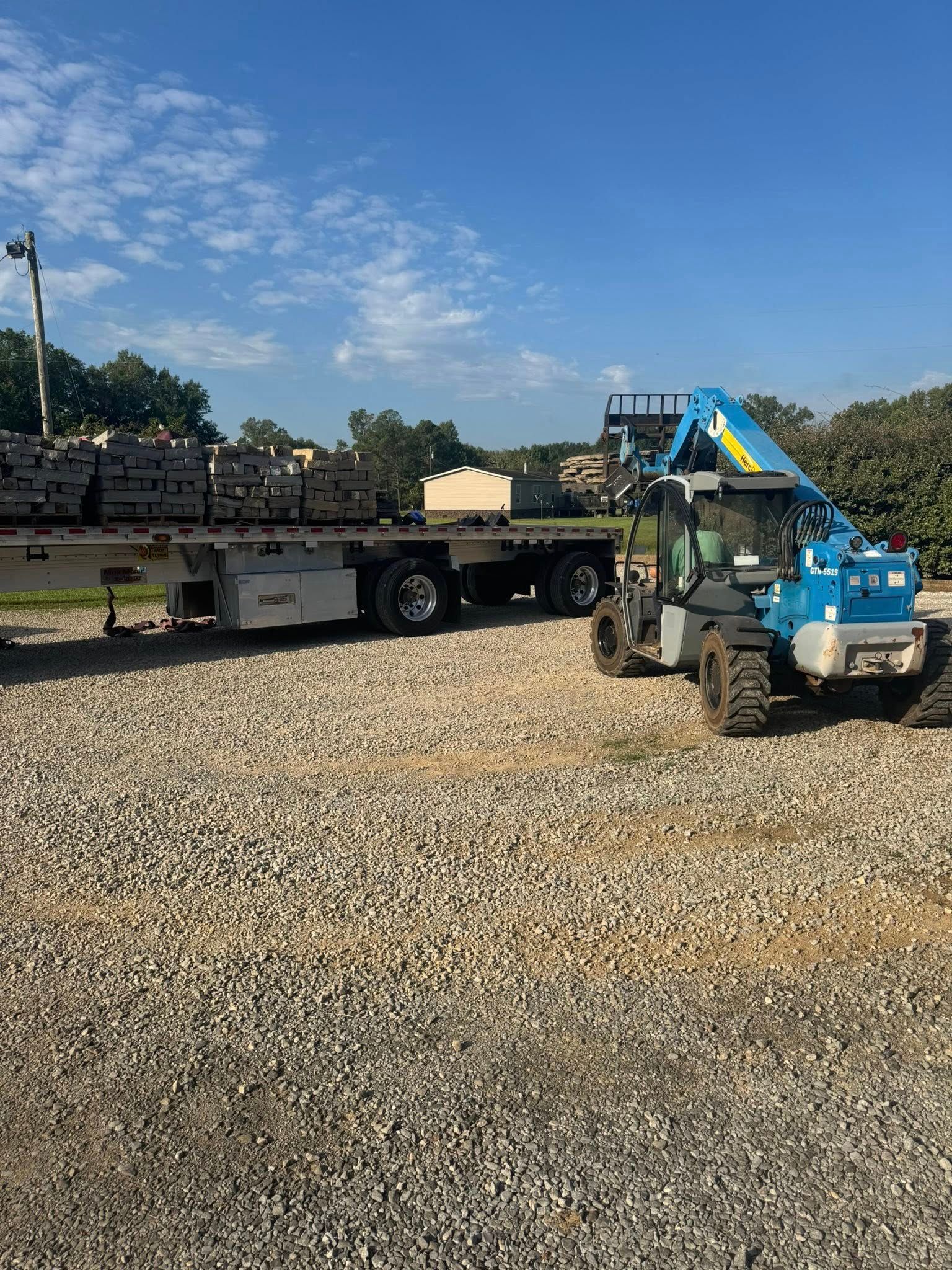Forklift unloading material from a flatbed trailer on a gravel lot under a blue sky.