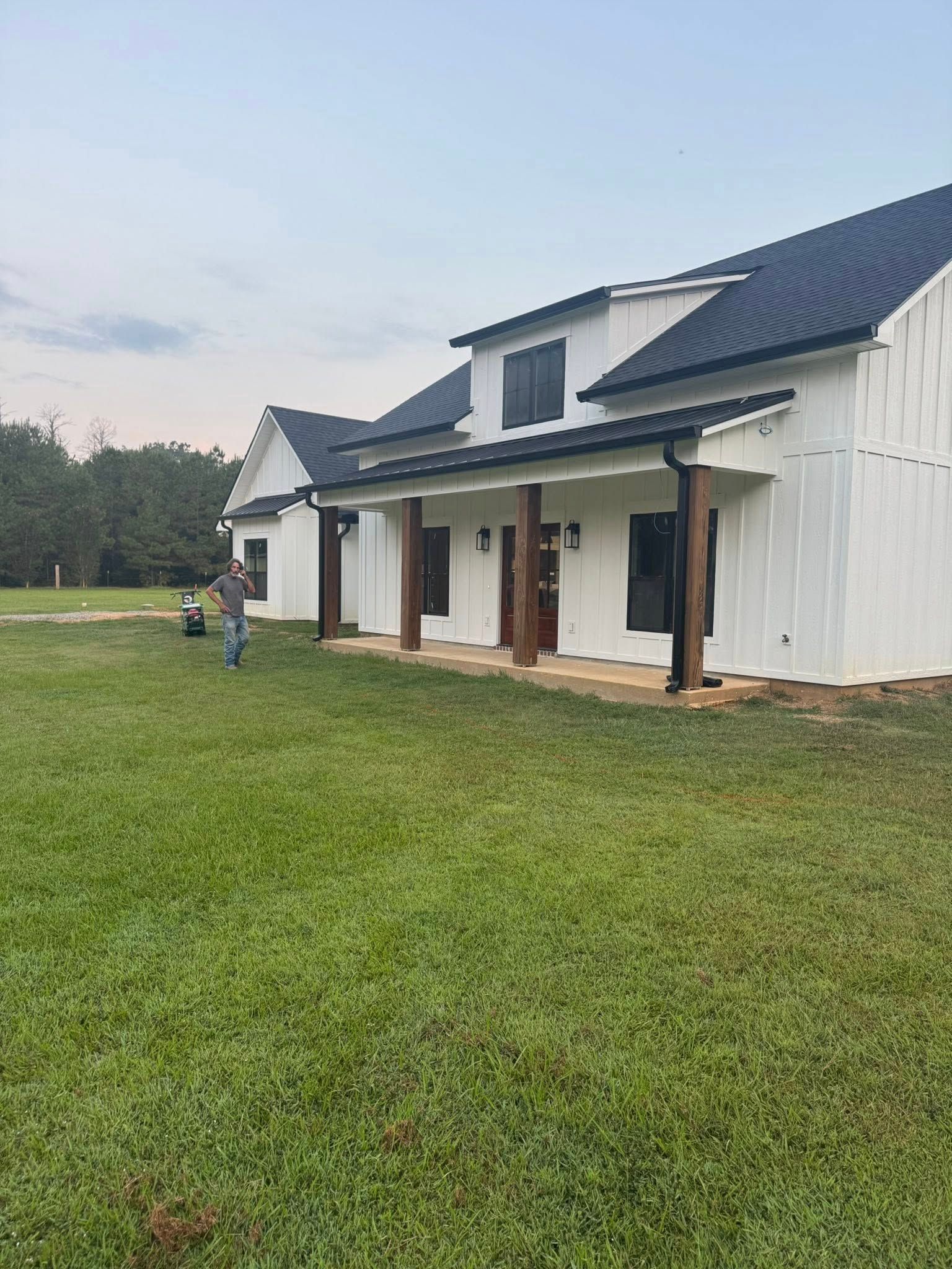White farmhouse with dark roof, brown columns, and a person mowing the lawn.