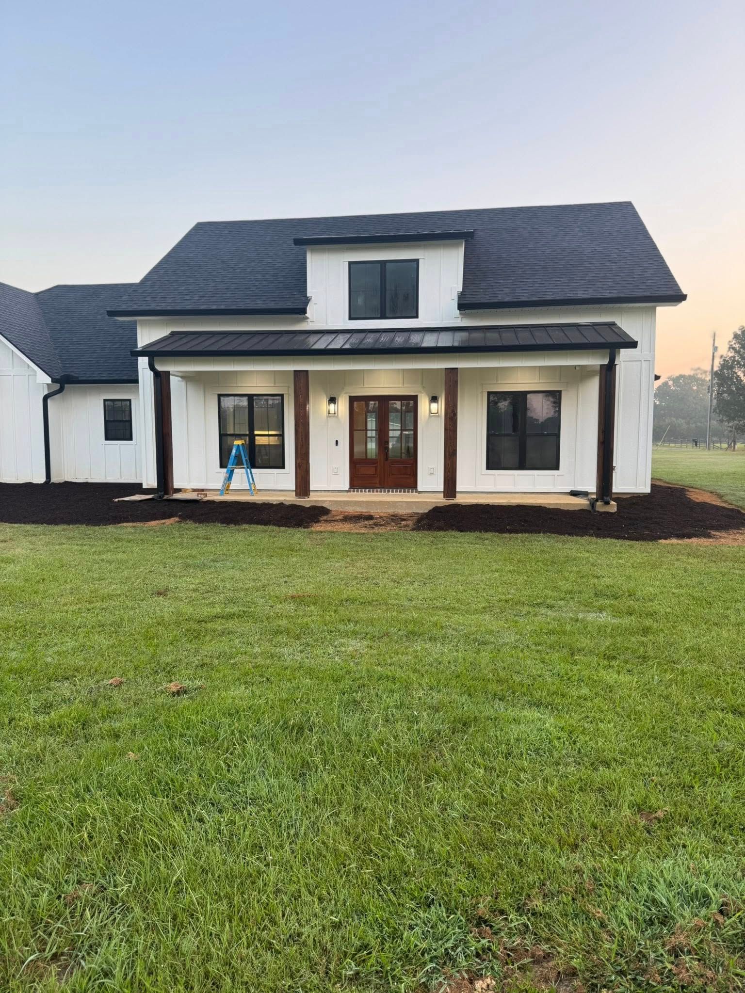 White farmhouse with dark roof and brown accents, porch with pillars, and green lawn.