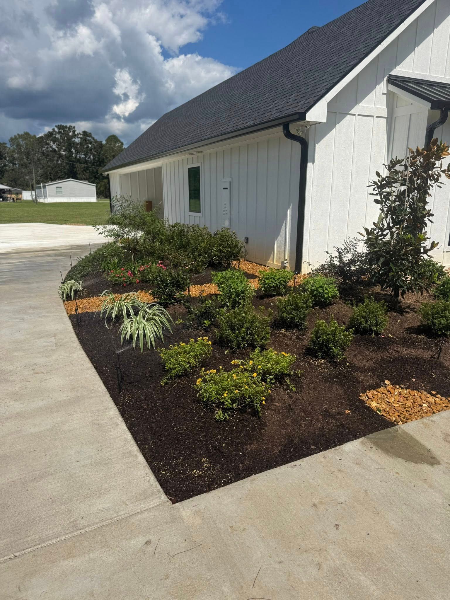White house with black roof, landscaped flower bed with mulch, concrete walkway, blue sky.