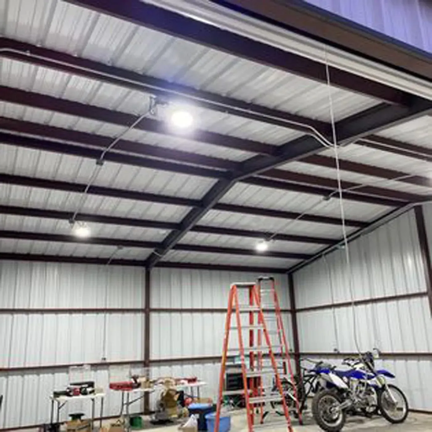 Interior of a metal building with overhead lighting, work table, tools, ladder, and a motorcycle.