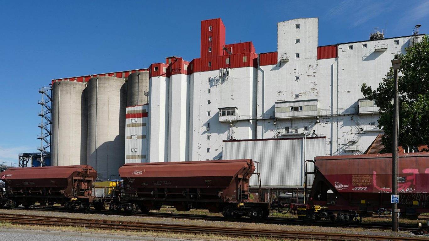 A freight train with reddish-brown hopper cars parked beside a large white and red industrial grain elevator facility.