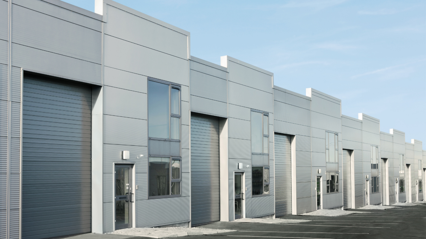 A row of modern, light gray industrial warehouse units featuring metal roller doors and tall vertical windows.