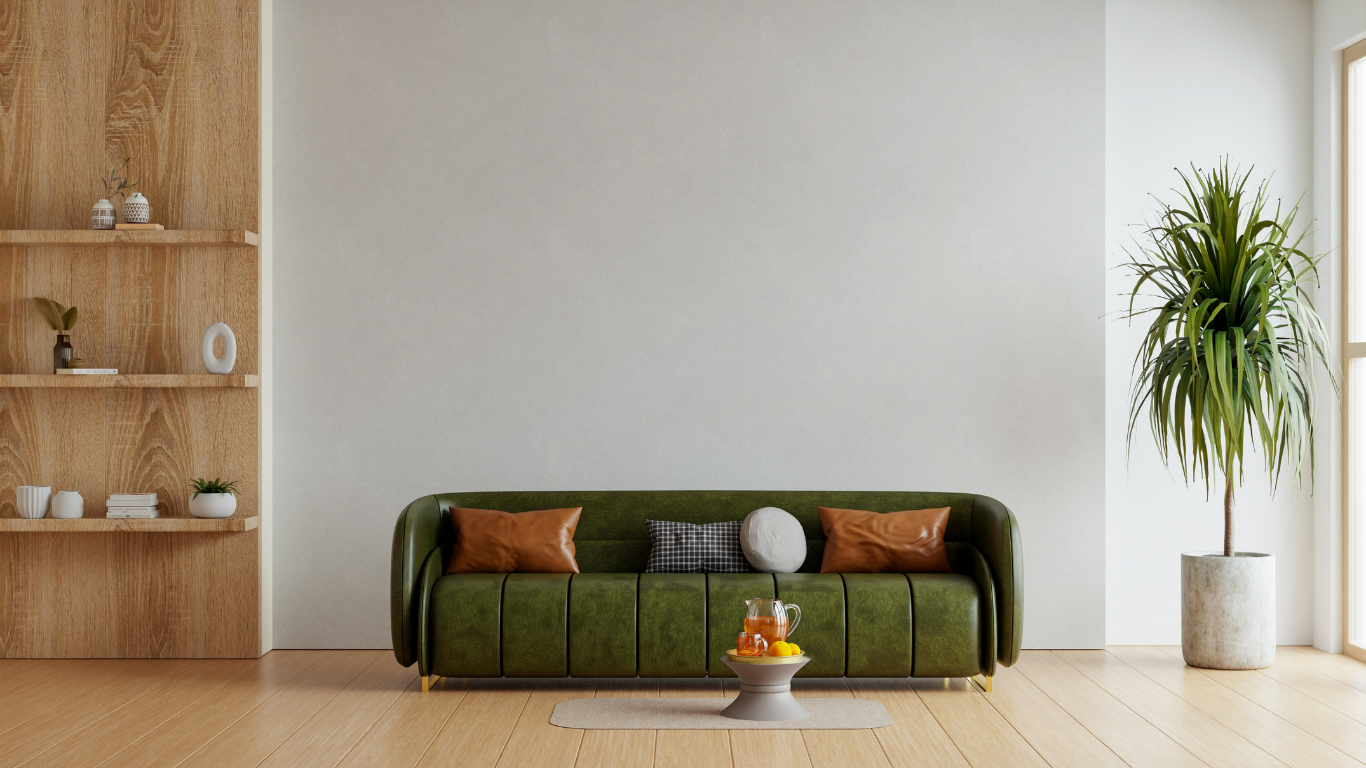 A green tufted sofa sits on a wooden floor against a plain wall, with a wooden shelf unit to the left and a tall plant.