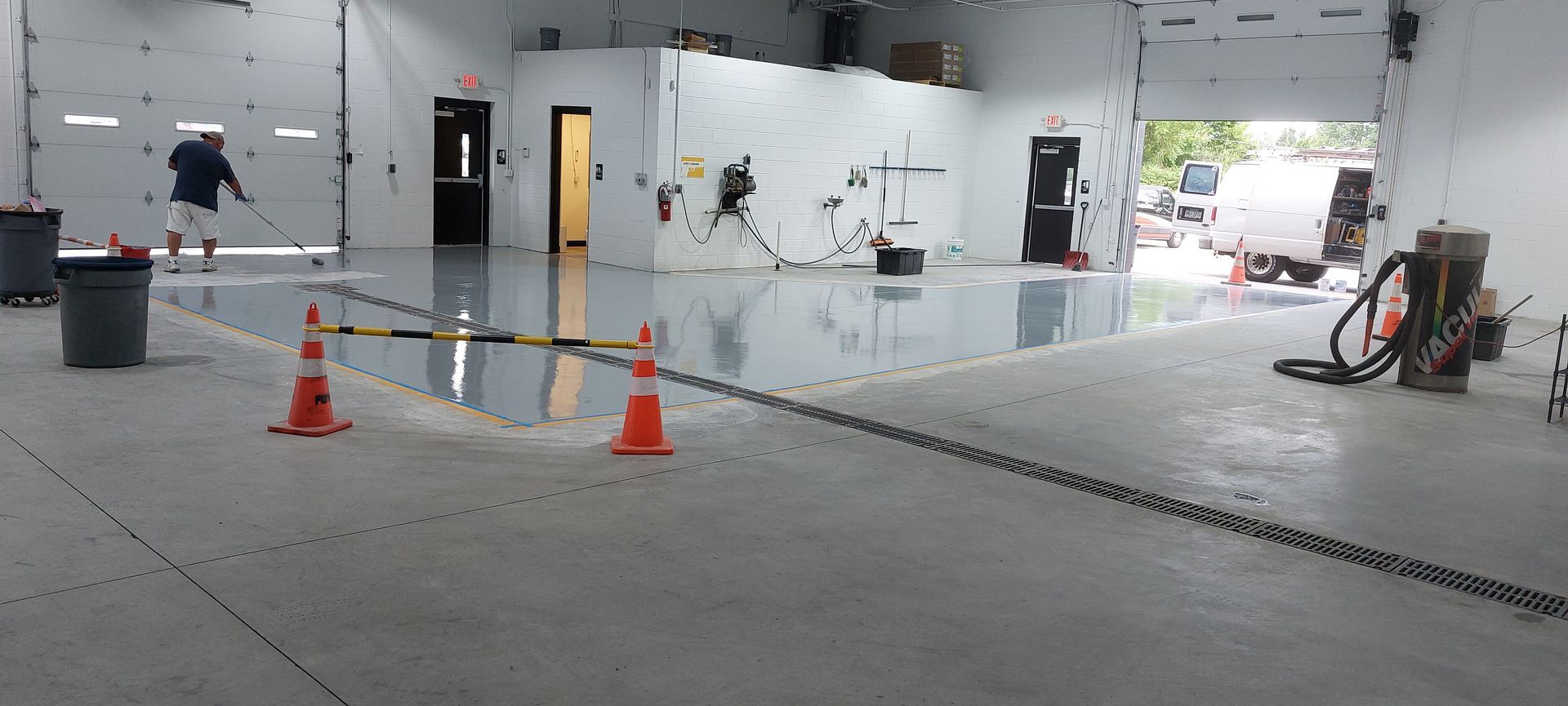 A person uses a squeegee to spread a glossy gray epoxy coating on a warehouse floor, with orange cones marking the area.