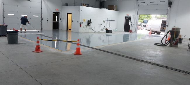 A person applies a smooth, reflective epoxy coating to a section of a concrete warehouse floor near two orange traffic cones.