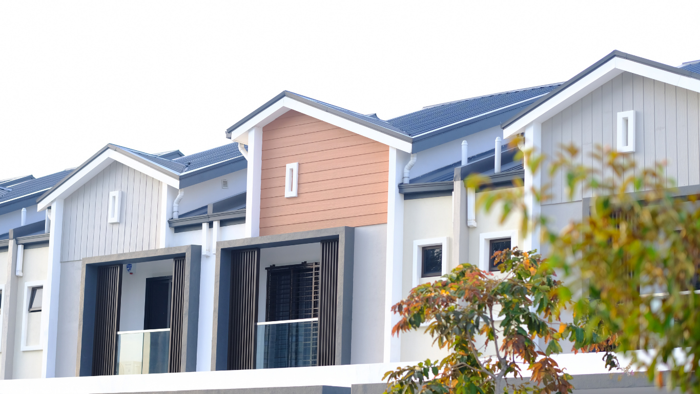 A row of modern two-story townhouses with gray and peach-toned facades, seen partially behind a leafy tree.