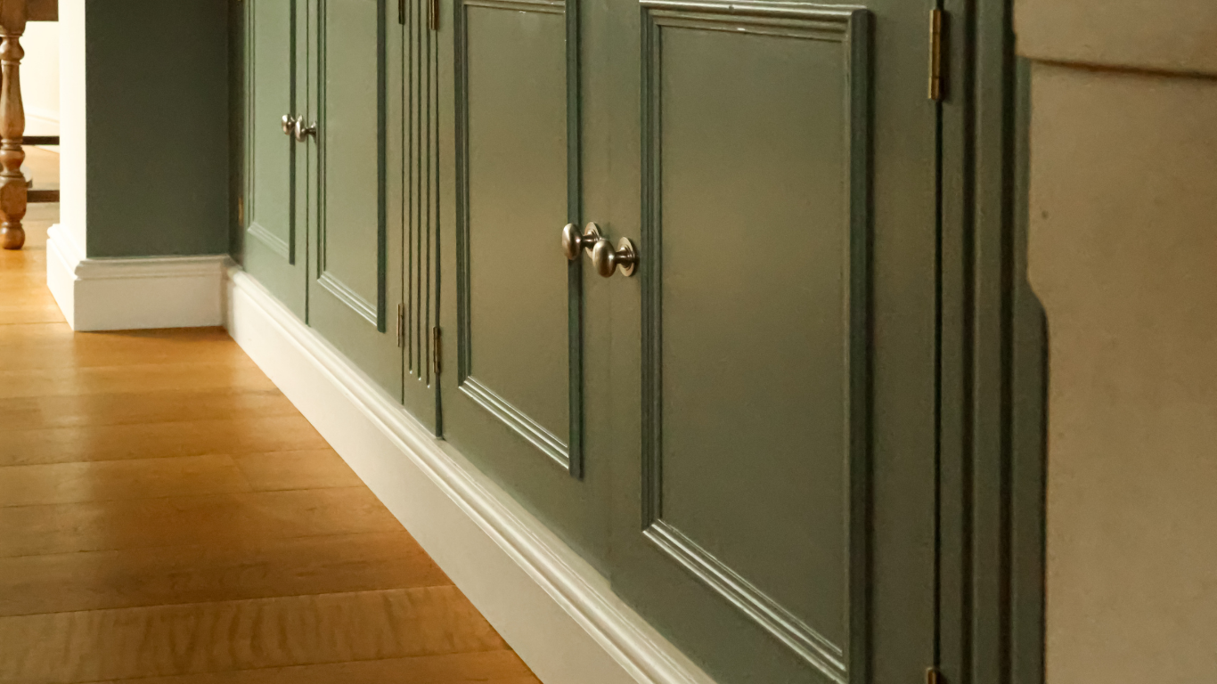 Sage green custom cabinets with brass knobs and white trim, set against a hardwood floor.