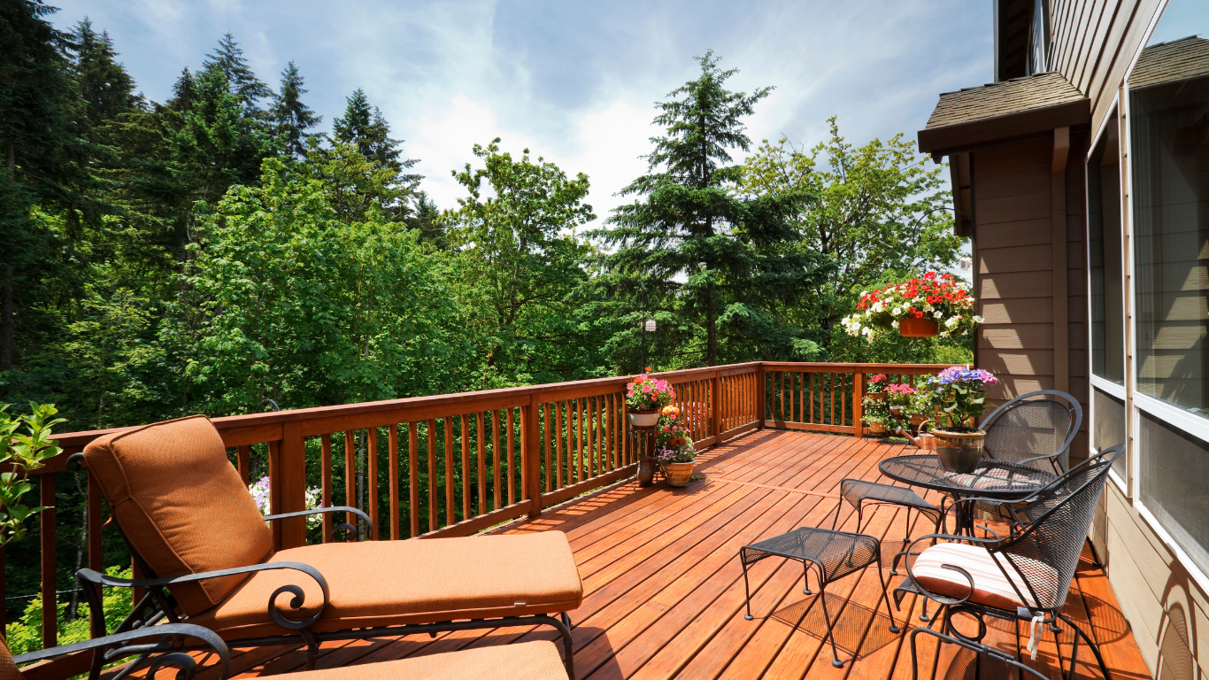 A wooden deck with a lounge chair, table, and chairs overlooking a lush, green forest on a sunny day.