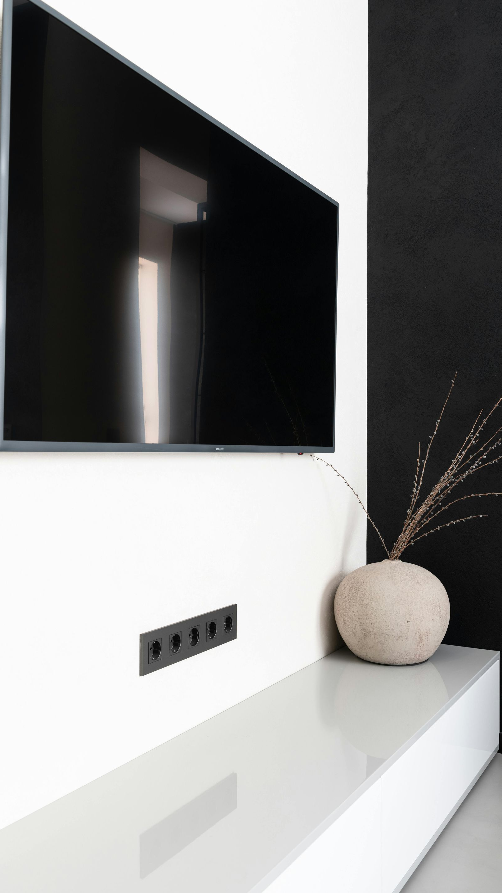 A wall-mounted TV above a long, white shelf with a small decorative vase and a row of black wall outlets.