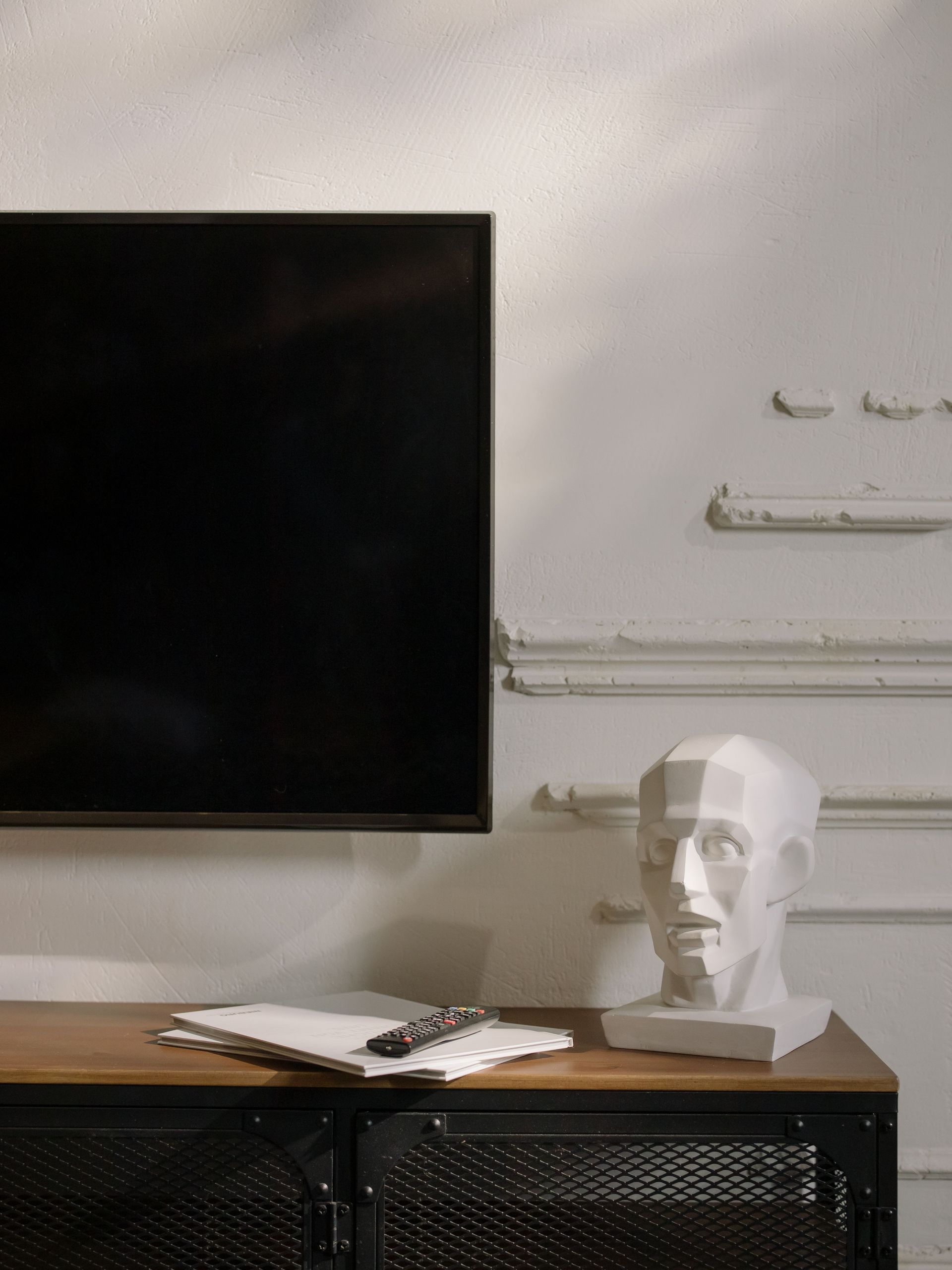 Wall-mounted black TV above a wooden desk with an open notebook and a white sculpted head