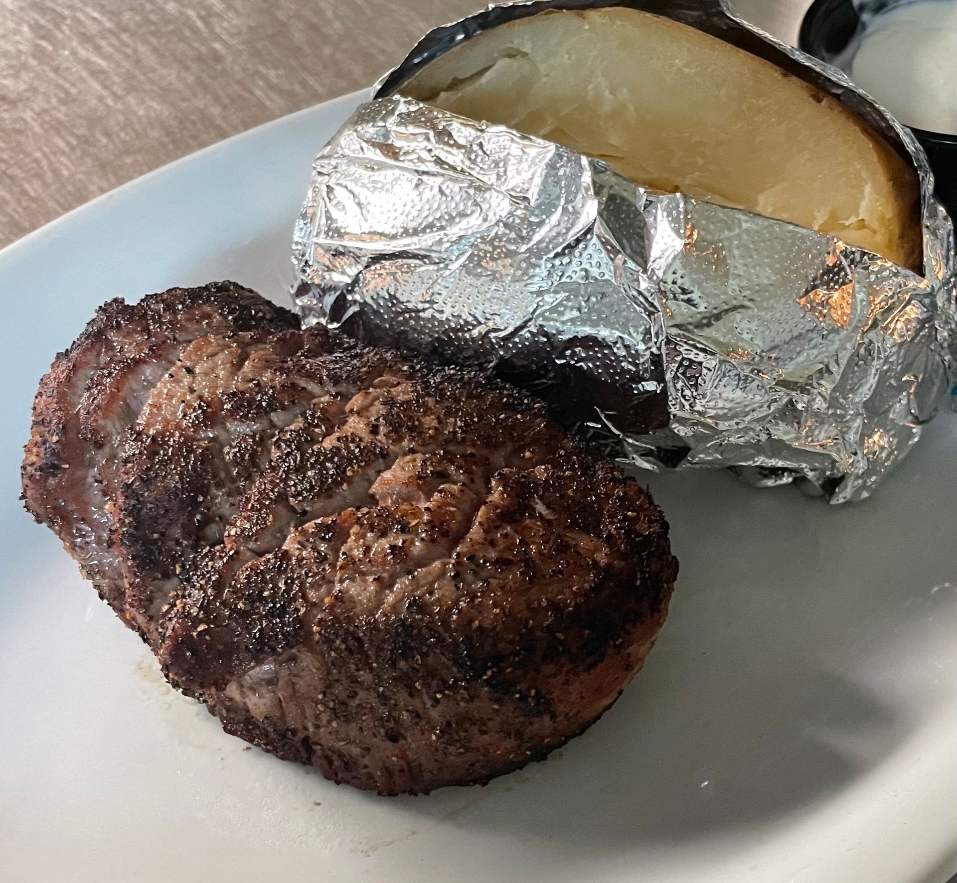 A white plate topped with a steak and a baked potato wrapped in tin foil