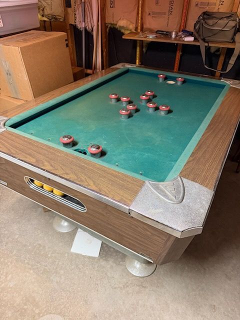 Coin-operated tabletop shuffleboard game in a garage, with metal pucks on a green playing surface.