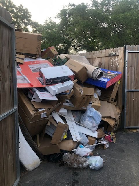 Overflowing dumpster piled with cardboard, trash, and debris beside a wooden fence outdoors