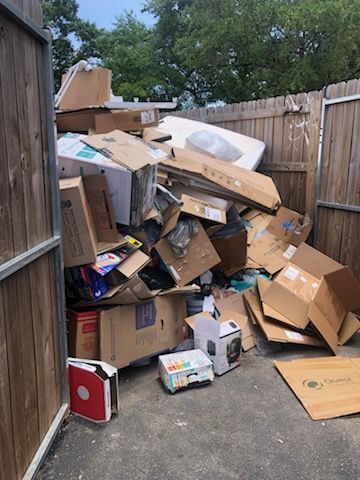Overflowing pile of cardboard boxes and debris stacked in a narrow outdoor alley by a wooden fence.