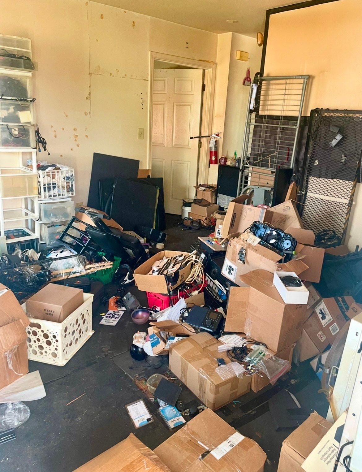 Cluttered room with many boxes, scattered items, and stacked storage bins near an open door.