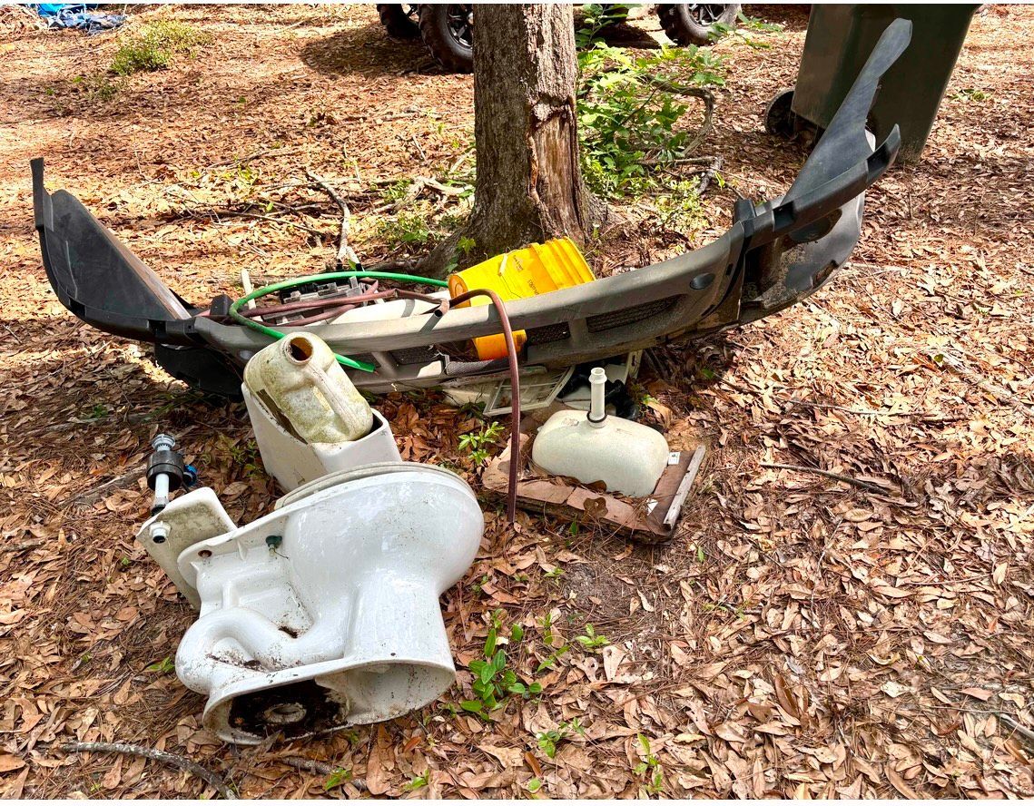 Broken white machinery and detached parts леж on leaf-covered ground beside a tree.
