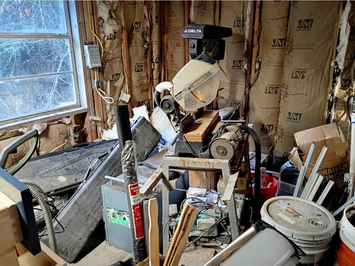 Cluttered workshop with a miter saw, wood scraps, tools, and construction materials by a window.