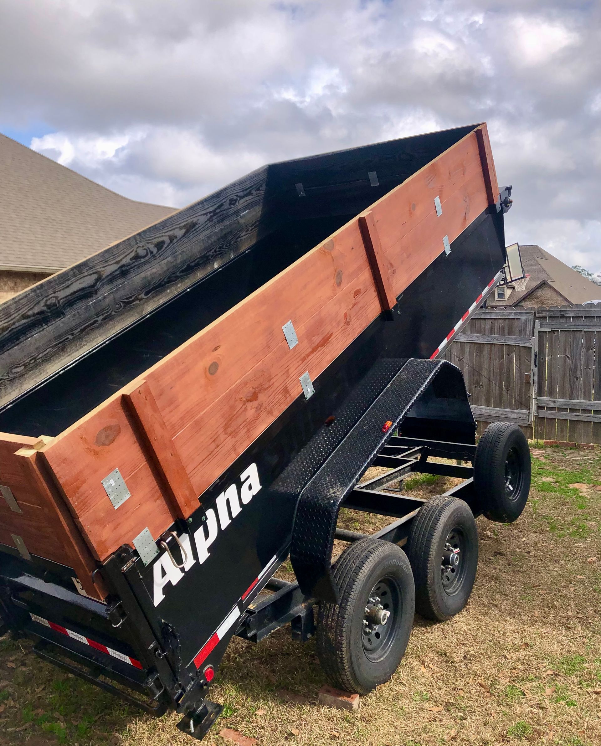 A black and orange dump trailer tipped steeply upward on a grassy lot under cloudy skies.