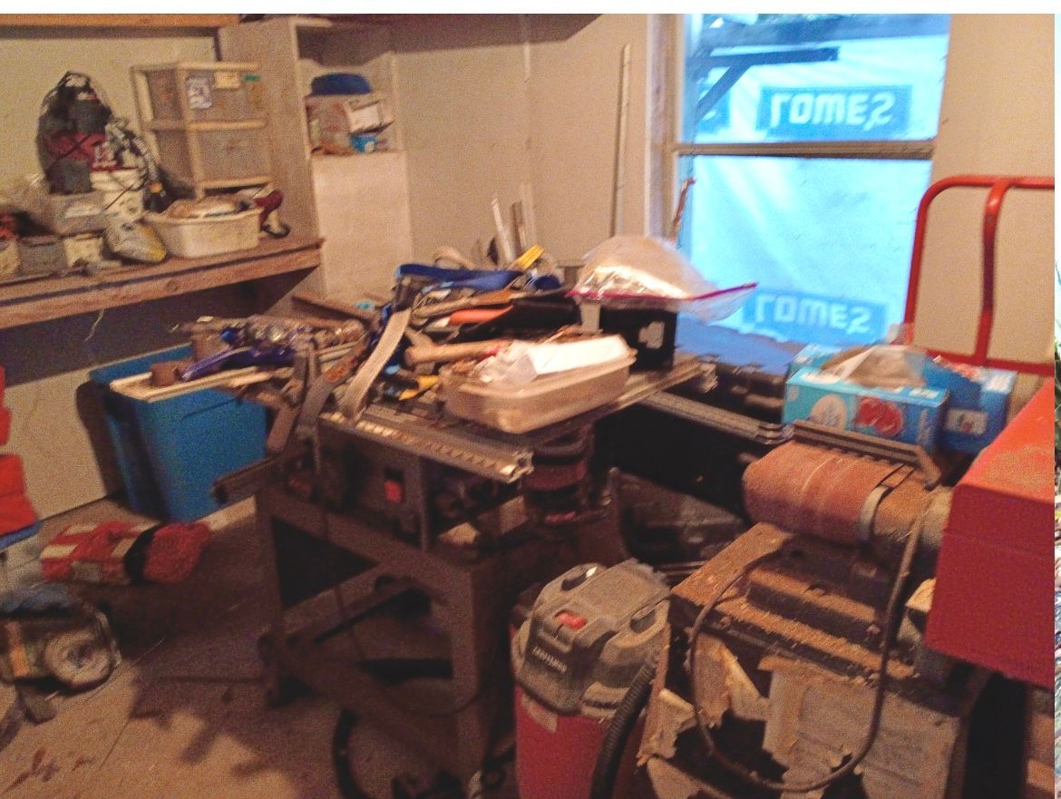Cluttered room with stacked boxes and tools, a workbench, and a glowing computer monitor in the corner