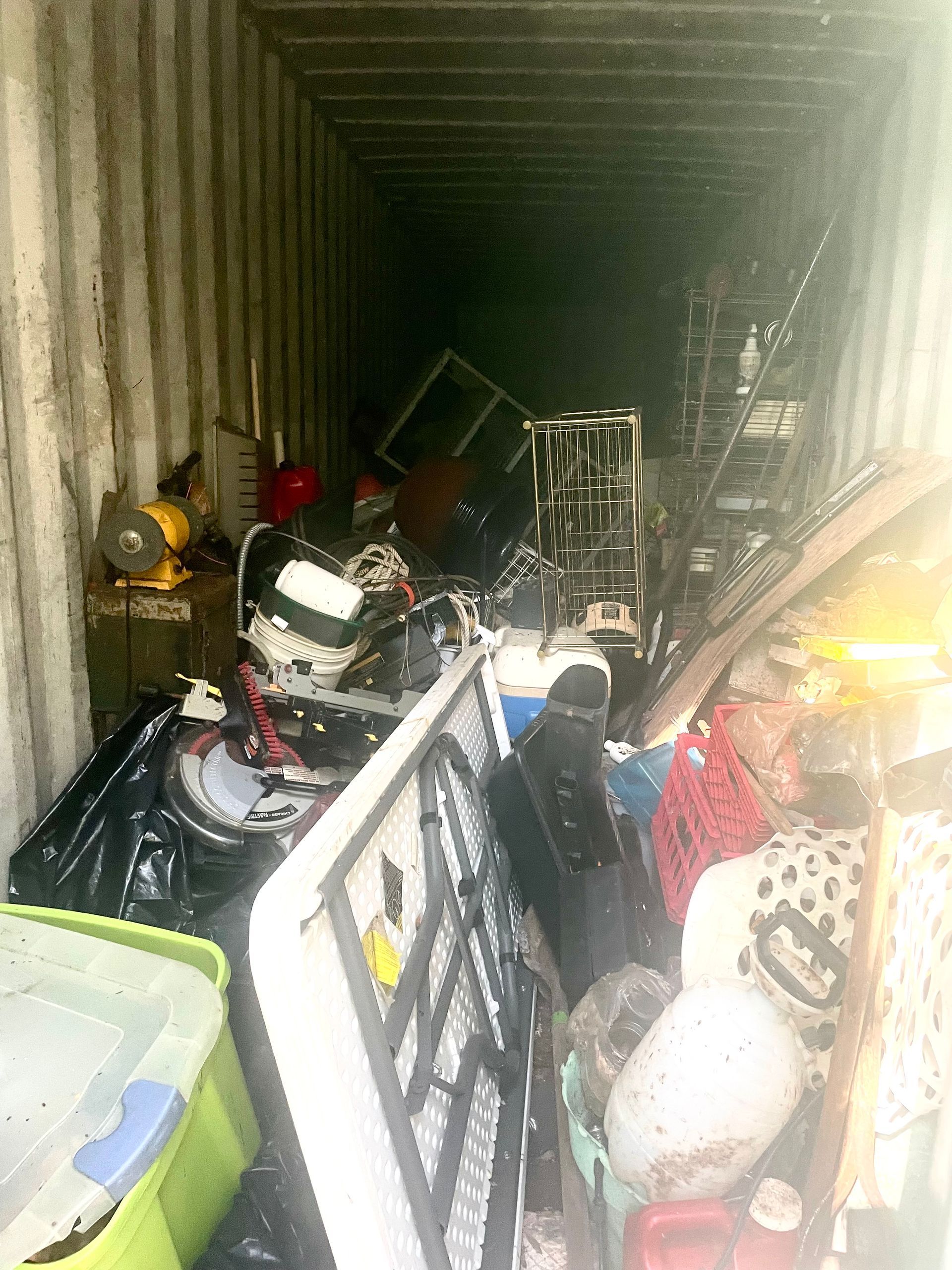 Cluttered shipping container packed with bags, bins, and metal frames, with bright light at the entrance