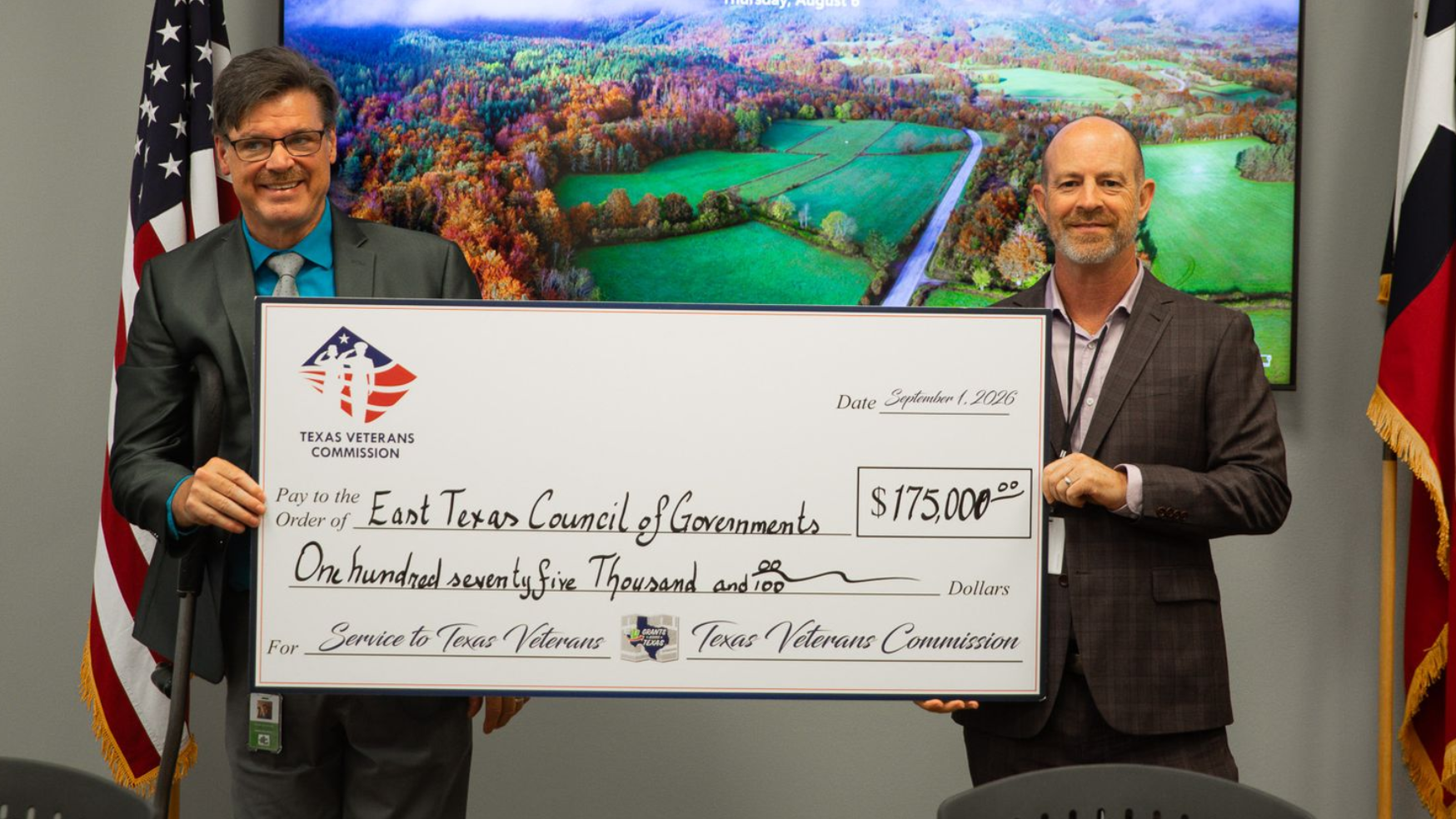 Two men holding a large ceremonial check in a conference room with a colorful aerial landscape display behind them